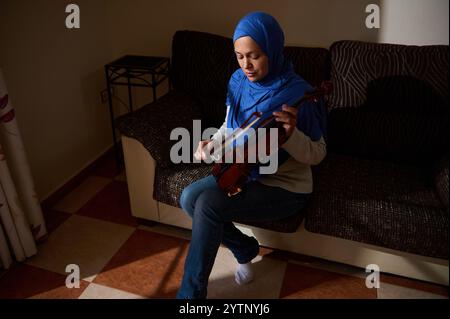 Une femme portant un hijab bleu joue du violon tout en étant assise sur un canapé, créant une atmosphère sereine et concentrée dans un cadre de salon confortable. Banque D'Images