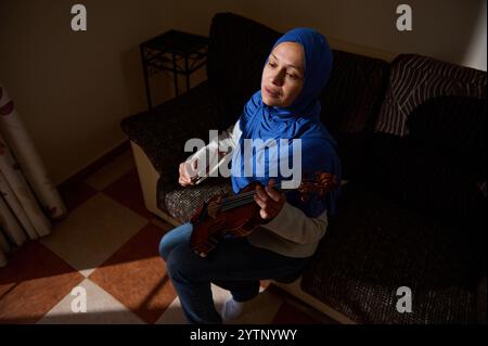 Une femme contemplative portant un hijab bleu joue du violon assise sur un canapé, baignée de lumière chaude. Banque D'Images