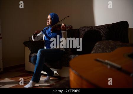 Une femme portant un hijab bleu joue du violon, confortablement assise sur un canapé dans une pièce chaleureusement éclairée, exprimant la sérénité et la passion pour la musique. Banque D'Images