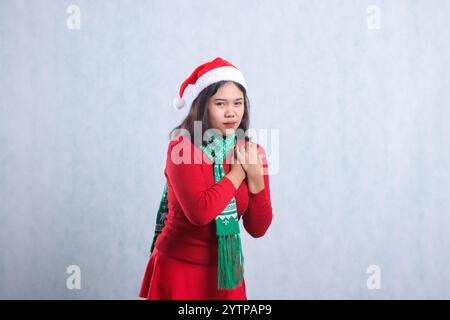 portrait de belle fille portant un pull de noël, chapeau de père noël et écharpe, maladie cardiaque du côté gauche regardant la caméra les deux mains tenant la poitrine, isoler Banque D'Images