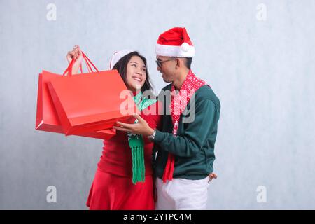 Charmant jeune couple portant des pulls de Noël rouges, des chapeaux de Père Noël et des foulards, joyeux se regardant les uns les autres soulevant des sacs cadeaux de Noël rouges, isolé Banque D'Images