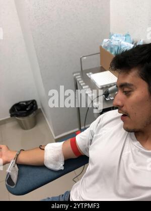 Un jeune homme est assis dans une chaise confortable dans une clinique de don de sang. Il porte un t-shirt blanc décontracté et a une bande autour de son bras pour se préparer Banque D'Images