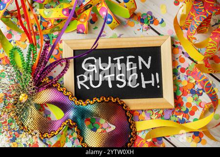 Bavière, Allemagne - 8 décembre 2024 : bonne année pour le réveillon du nouvel an sur un tableau noir, entouré de banderoles colorées, de confettis et d'un masque de fête. PHOTOMONTAGE *** Guten Rutsch Gruß zu Silvester auf einer Tafel, umgeben von bunten Luftschlangen, Konfetti und einer festlichen Partymaske. FOTOMONTAGE Banque D'Images