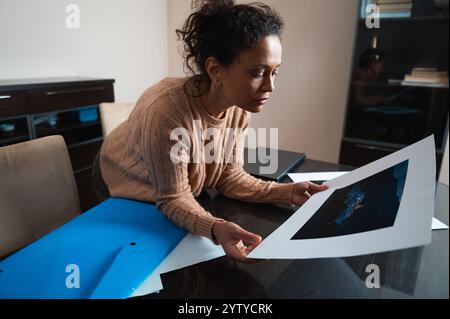 Une femme en pull beige examine attentivement une œuvre d'art dans un bureau à domicile moderne et confortable, mettant en valeur la créativité et la concentration. Banque D'Images