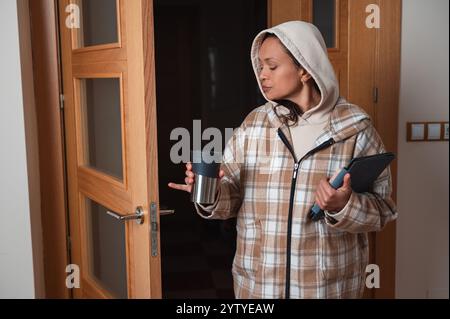 Une femme dans un manteau à carreaux et un sweat à capuche tient une tablette et une tasse tout en fermant une porte, suggérant une routine matinale confortable. Banque D'Images