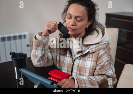 Une femme réfléchie en tenue décontractée tient une carte de crédit tout en utilisant une tablette. Un portefeuille rouge et un gobelet noir sont visibles sur la table, représentant un M. Banque D'Images