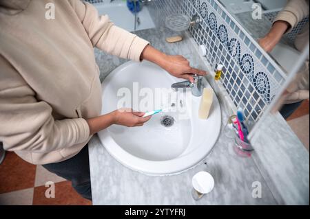 Une personne dans un sweat à capuche beige nettoie une brosse à dents sous le robinet d'un évier en marbre élégant. Banque D'Images