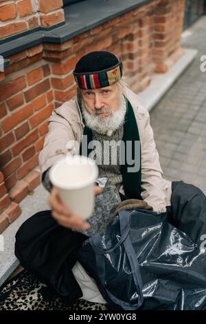 Vue à angle bas d'un homme âgé sans abri affamé dans des vêtements portés assis sur la rue urbaine et mendiant pour l'aumône, tenant une tasse jetable. Banque D'Images