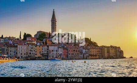 Alors que le soleil plonge sous l'horizon, le front de mer de Rovinj Croatie prend vie avec des teintes chaudes, mettant en valeur des bâtiments colorés, une église imposante et tranqui Banque D'Images