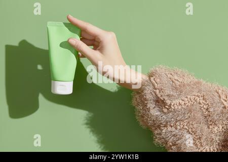 Une main de femme dans un pull tient un tube de crème sur un fond vert. Soins de beauté auto-soignés Banque D'Images