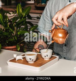 Une personne versant élégamment du thé parfumé d'une superbe théière dorée dans des tasses délicates au milieu d'une végétation luxuriante Banque D'Images