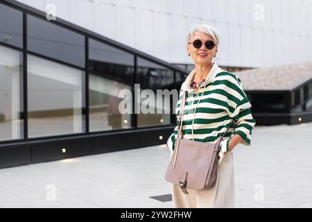 Portrait belle femme mature heureuse dans une tenue élégante à l'extérieur dans la ville avec une coiffure courte portant un pull rayé et des lunettes de soleil, regardant C Banque D'Images