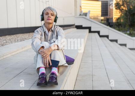 Réfléchie solitaire femme mature à la mode écoutant de la musique sur des écouteurs tout en étant assis sur les marches de White Modern Building dans la ville. Banque D'Images