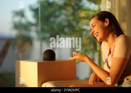 Femme heureuse parlant à haut-parleur intelligent au coucher du soleil à la maison Banque D'Images