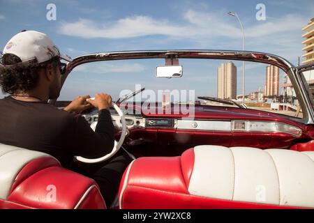 Homme conduisant une voiture cabriolet d'époque avec une vue d'un paysage urbain en arrière-plan, la Havane, Cuba Banque D'Images