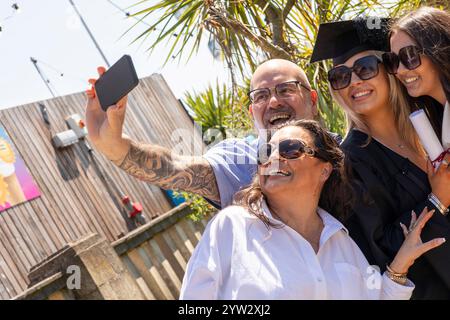 Un groupe joyeux prend un selfie pour célébrer une remise des diplômes, avec deux femmes souriantes portant des casquettes de remise des diplômes et un homme tatoué portant des lunettes, Bournemouth, Dorset UK Banque D'Images