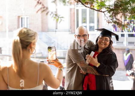 Fier homme embrassant une diplômée féminine souriante en casquette et robe tandis qu'une autre personne prend sa photo avec un smartphone, Bournemouth, Dorset UK Banque D'Images