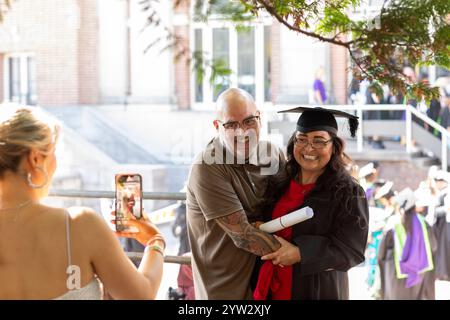 Célébration du jour de la remise des diplômes avec un homme fier embrassant une femme en casquette et robe alors qu'une autre personne prend sa photo, Bournemouth, Dorset UK Banque D'Images