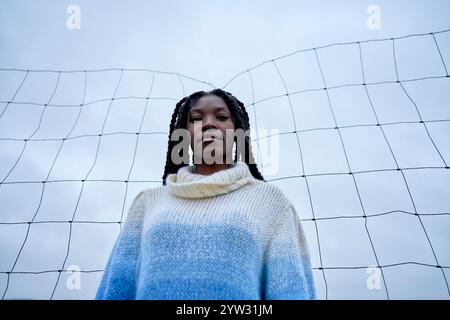 Jeune femme debout devant une structure de filet sous un ciel nuageux, regardant attentivement le spectateur, Brandebourg, Allemagne Banque D'Images