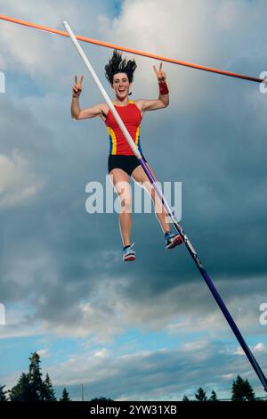 Voûte à perche féminine en plein air au-dessus de la barre contre un fond de ciel nuageux, arborant des vêtements athlétiques lumineux et une expression joyeuse. Banque D'Images