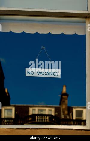 Un panneau No Vacancy est accroché à une fenêtre contre un ciel bleu clair, reflétant les bâtiments et un cadre de fenêtre rideau, Édimbourg, Écosse Banque D'Images