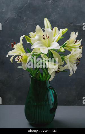 Élégants lis blancs disposés dans un vase en verre vert sur un fond texturé foncé. Composition florale minimaliste mettant l'accent sur la simplicité et la beauté Banque D'Images