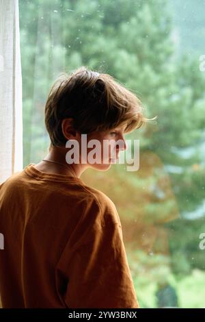 Jeune personne regardant par une fenêtre un jour de pluie, avec des gouttes visibles sur le verre et de la verdure en arrière-plan, Norvège Banque D'Images