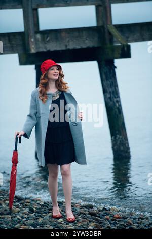 Une femme vibrante dans un chapeau rouge se tient près d'un rivage brumeux, tenant un parapluie rouge, avec une jetée en bois en arrière-plan. Banque D'Images