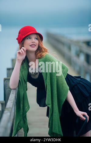 Jeune femme souriante aux cheveux roux portant un chapeau rouge et un châle vert appuyé sur une balustrade en bois près de l'eau. Banque D'Images