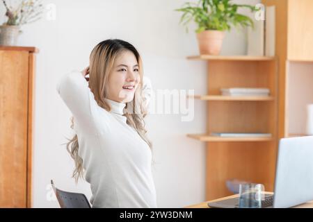 Femme souriante avec de longs cheveux ondulés assis à un bureau avec un ordinateur portable, s'étirant avec les mains derrière la tête dans une pièce bien éclairée. Banque D'Images