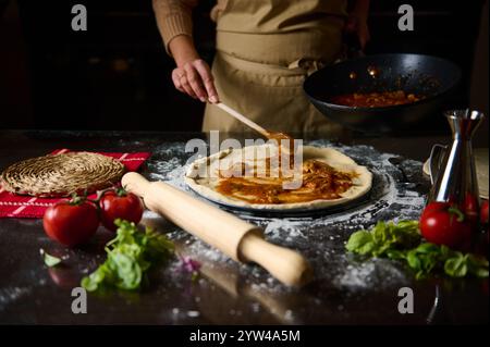 Une personne dans un tablier étale de la sauce tomate sur de la pâte à pizza fraîche. Les ingrédients environnants comprennent des tomates et des herbes, créant un atmos confortable et fait maison Banque D'Images