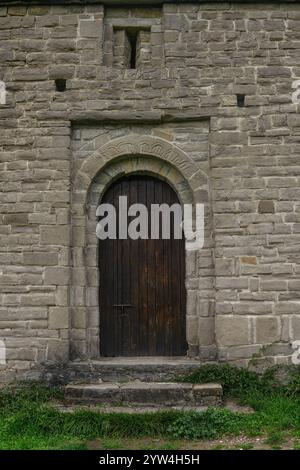 Chapelle en pierre au design rustique, située dans un champ verdoyant avec un fond montagneux sous un ciel nuageux. Banque D'Images