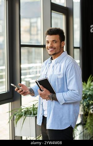 Un homme confiant se tient près de grandes fenêtres, souriant tout en tenant un téléphone et un ordinateur portable. Banque D'Images