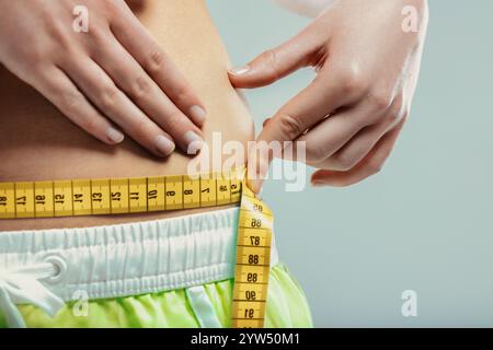 Gros plan d'une femme pinçant sa graisse du ventre tout en mesurant sa taille avec un ruban à mesurer jaune, exprimant des inquiétudes au sujet de la prise de poids et de l'image corporelle Banque D'Images