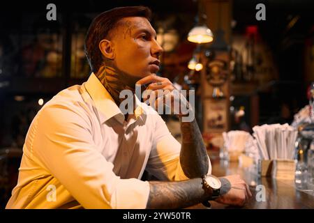 Un jeune homme avec des tatouages frappants est assis avec soin dans un bar, embrassant l'ambiance du soir. Banque D'Images