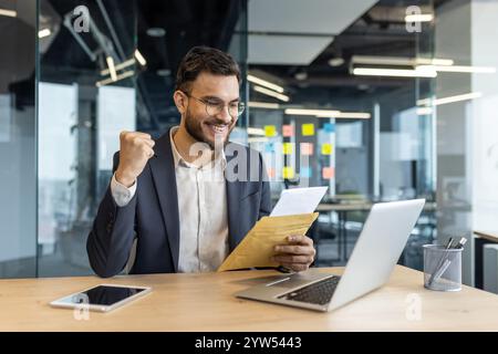 Un homme d'affaires heureux fête après avoir lu de bonnes nouvelles dans un bureau. Il tient une lettre et affiche de la joie, affichant le succès et l'optimisme. Un ordinateur portable et un smartphone sont visibles sur le bureau. Banque D'Images