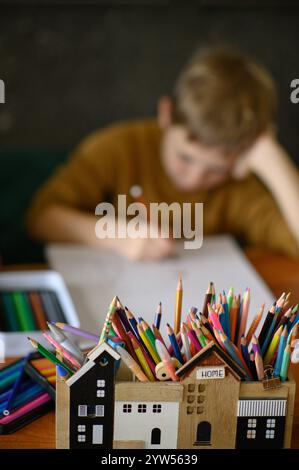 Petit garçon dessine une image d'hiver sur la table en bois et a des marqueurs et des crayons autour Banque D'Images