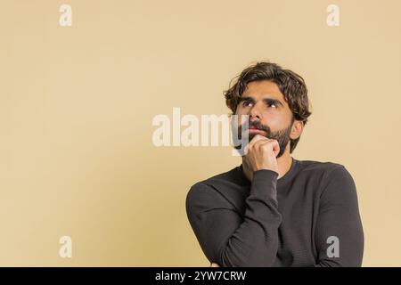 Réfléchi intelligent bel homme indien à manches longues regardant de côté avec une expression positive réfléchissant à une solution douteuse question. Homme adulte hispanique avec fond de studio beige. Les modes de vie des gens. Banque D'Images