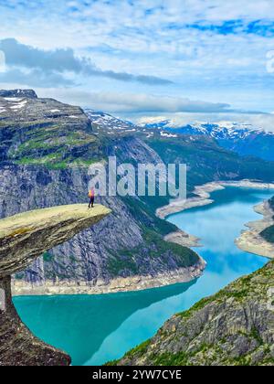 Un randonneur solitaire se tient au bord d'une falaise à couper le souffle, contemplant les eaux turquoises d'un fjord entouré de montagnes majestueuses en Norvège. Trolltu Banque D'Images