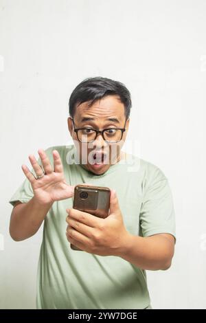 Portrait de l'homme asiatique surpris avec des lunettes regardant ce qu'il voit sur son smartphone sur fond blanc de mur de studio Banque D'Images
