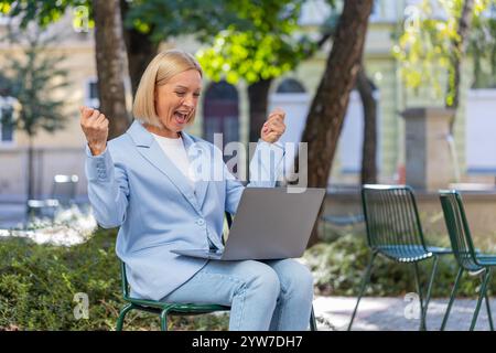 Femme d'affaires mature caucasienne excitée lisant de grandes nouvelles sur l'écran d'ordinateur portable tout en étant assise sur le banc sur la rue de la ville. Employeur féminin joyeux souriant en costume formel célébrant le succès dans les affaires Banque D'Images