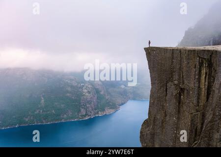 Une figure solitaire se dresse au bord de Preikestolen, une falaise spectaculaire en Norvège, surplombant une vaste étendue d'eau enveloppée de brume. Preikestolen, Norw Banque D'Images