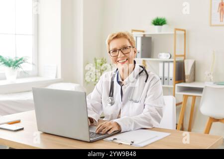 Médecin mature souriant assis au bureau travaillant dans le cabinet médical et regardant joyeux à la caméra. Banque D'Images
