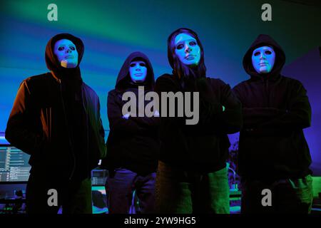 Portrait à angle bas du groupe de hackers portant un sweat à capuche noir et un masque blanc pour cacher leurs visages debout dans la chambre illumed avec des néons Banque D'Images