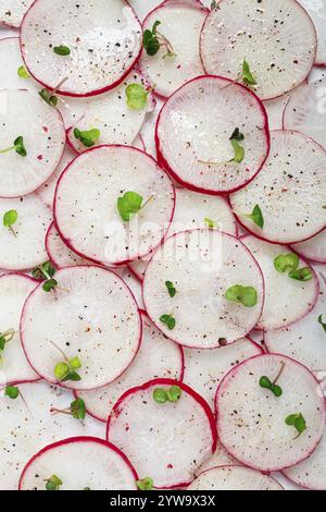 Des radis finement tranchés dans des couleurs vives rouge et blanc sont disposés sur une surface blanche, saupoudrés de poivre noir et ornés de petits microverts Banque D'Images