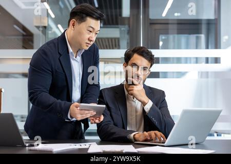 Deux professionnels s’engagent dans une discussion à l’aide d’un ordinateur portable et d’un smartphone dans un environnement de bureau moderne, mettant l’accent sur la collaboration et la planification stratégique. Banque D'Images