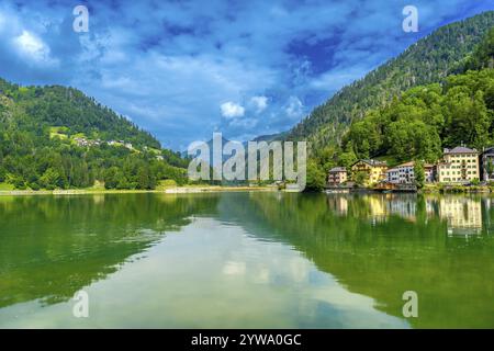 Scène estivale tranquille au lac alleghe avec des maisons colorées reflétées dans l'eau émeraude, entouré de montagnes verdoyantes sous un ciel bleu vibrant Banque D'Images