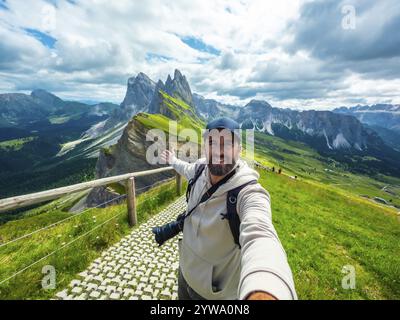 Photographe avec sac à dos et appareil photo prenant un selfie avec le bras tendu avec le groupe de montagne odle di eores en arrière-plan Banque D'Images