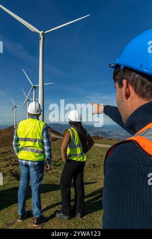 Trois ingénieurs multiethniques debout sur une colline, observant des éoliennes générant de l’énergie propre, tout en discutant de l’avancement du projet et du prosp futur Banque D'Images