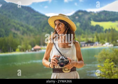 Femme portant un chapeau de paille tenant un appareil photo vintage profitant d'une vue imprenable sur le lac dobbiaco et les montagnes environnantes dans les dolomites Banque D'Images